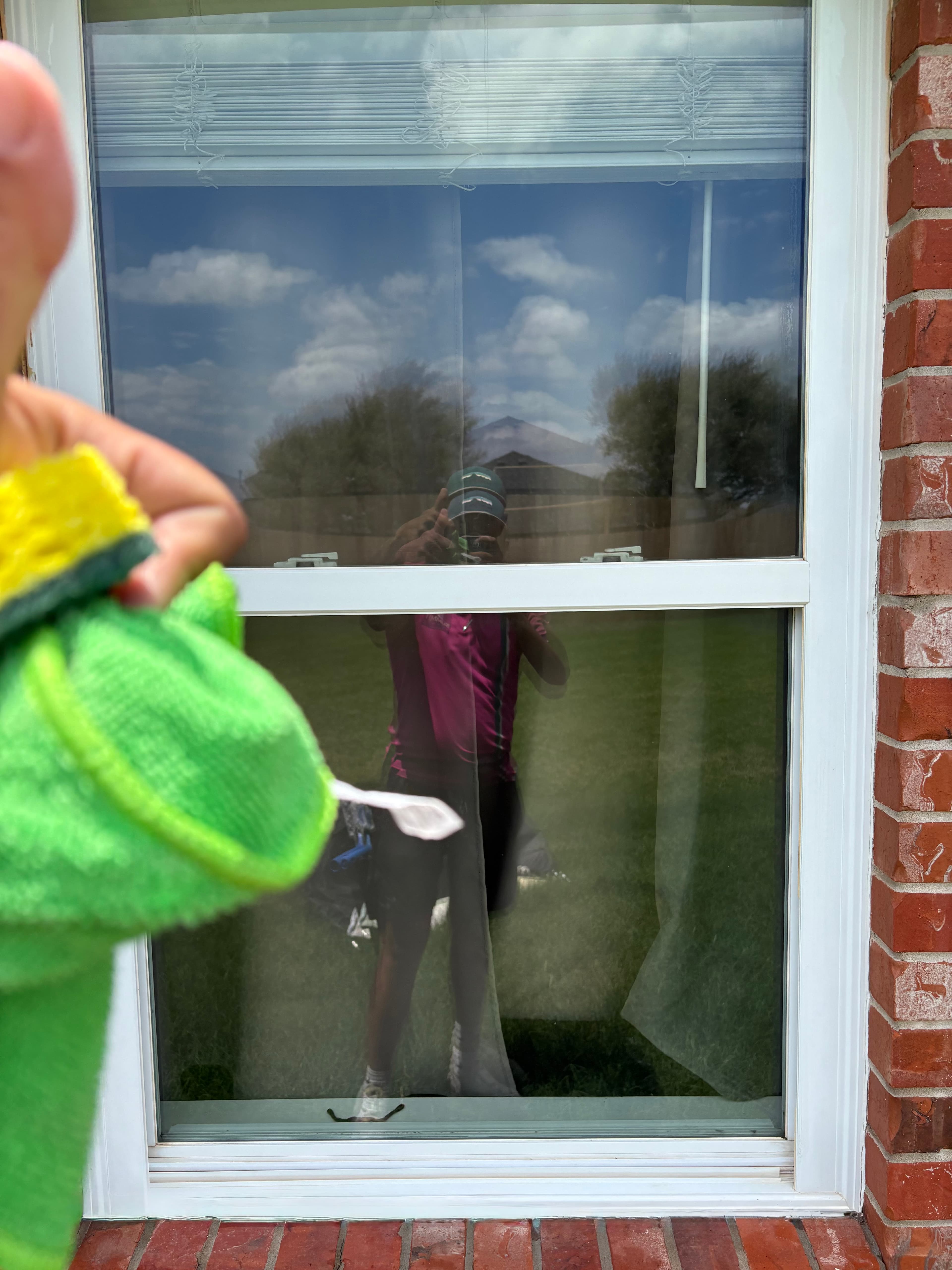 Hand holding a green sponge near a window reflecting a person taking a picture.