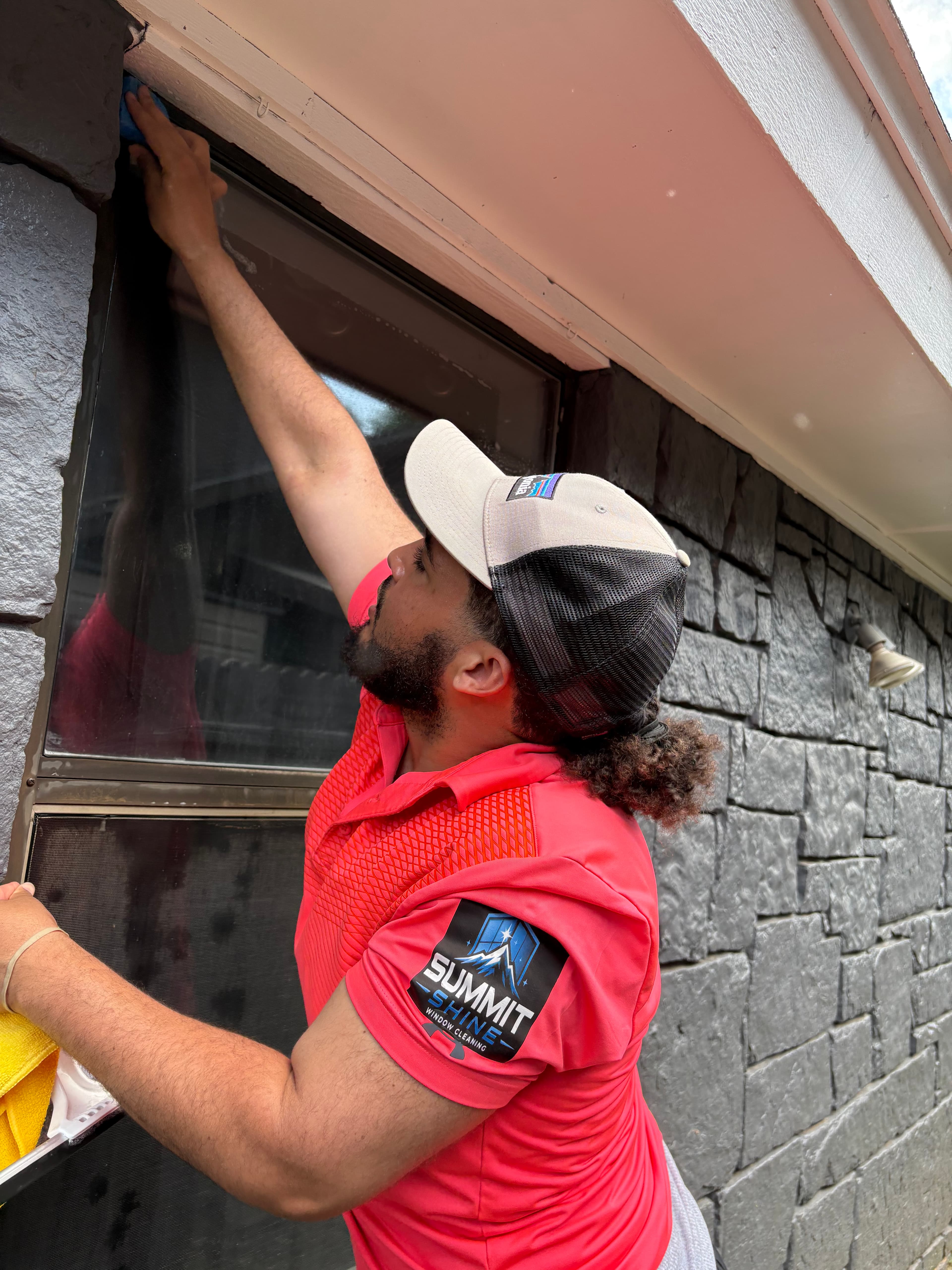 Man in a red polo shirt cleaning the top corner of an exterior window frame.