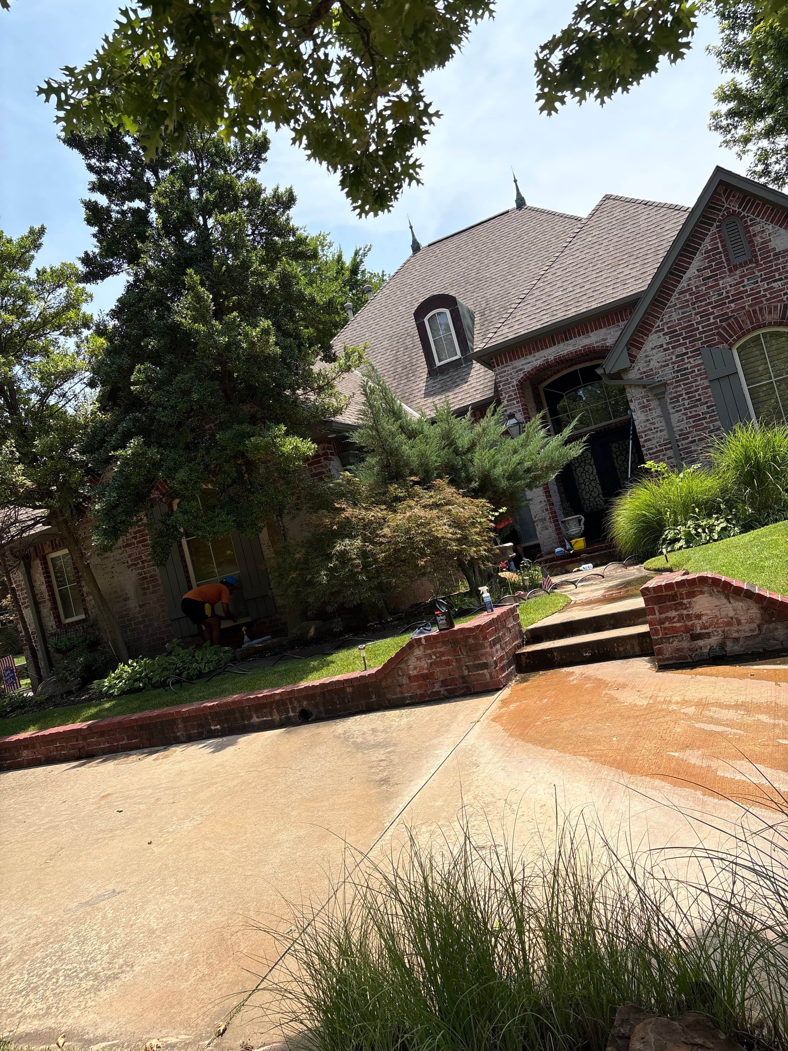 Large brick house with steep rooflines and lush green landscaping bordering a concrete driveway.