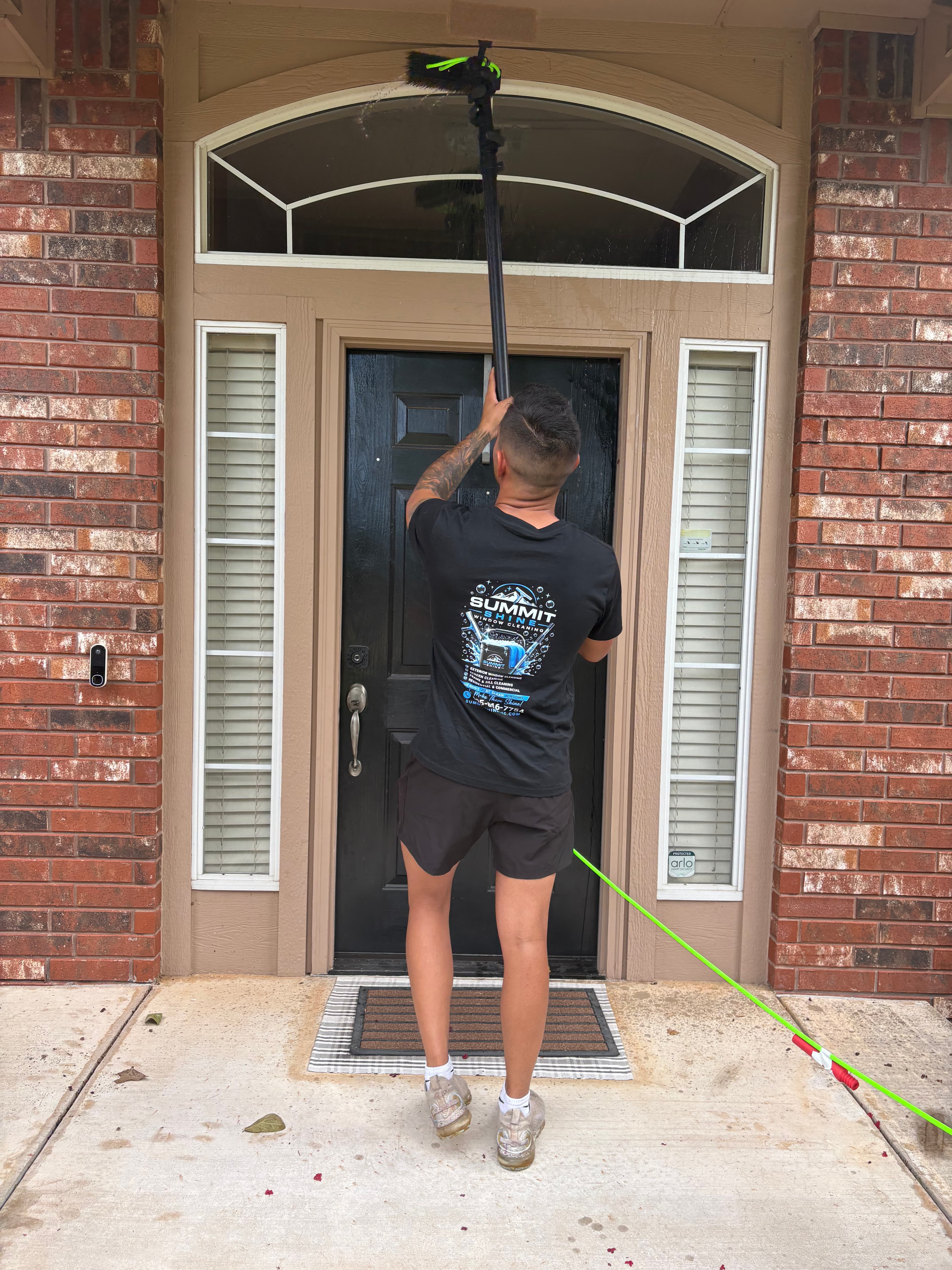 Man cleaning an arched window above a black front door with a long-reach brush.