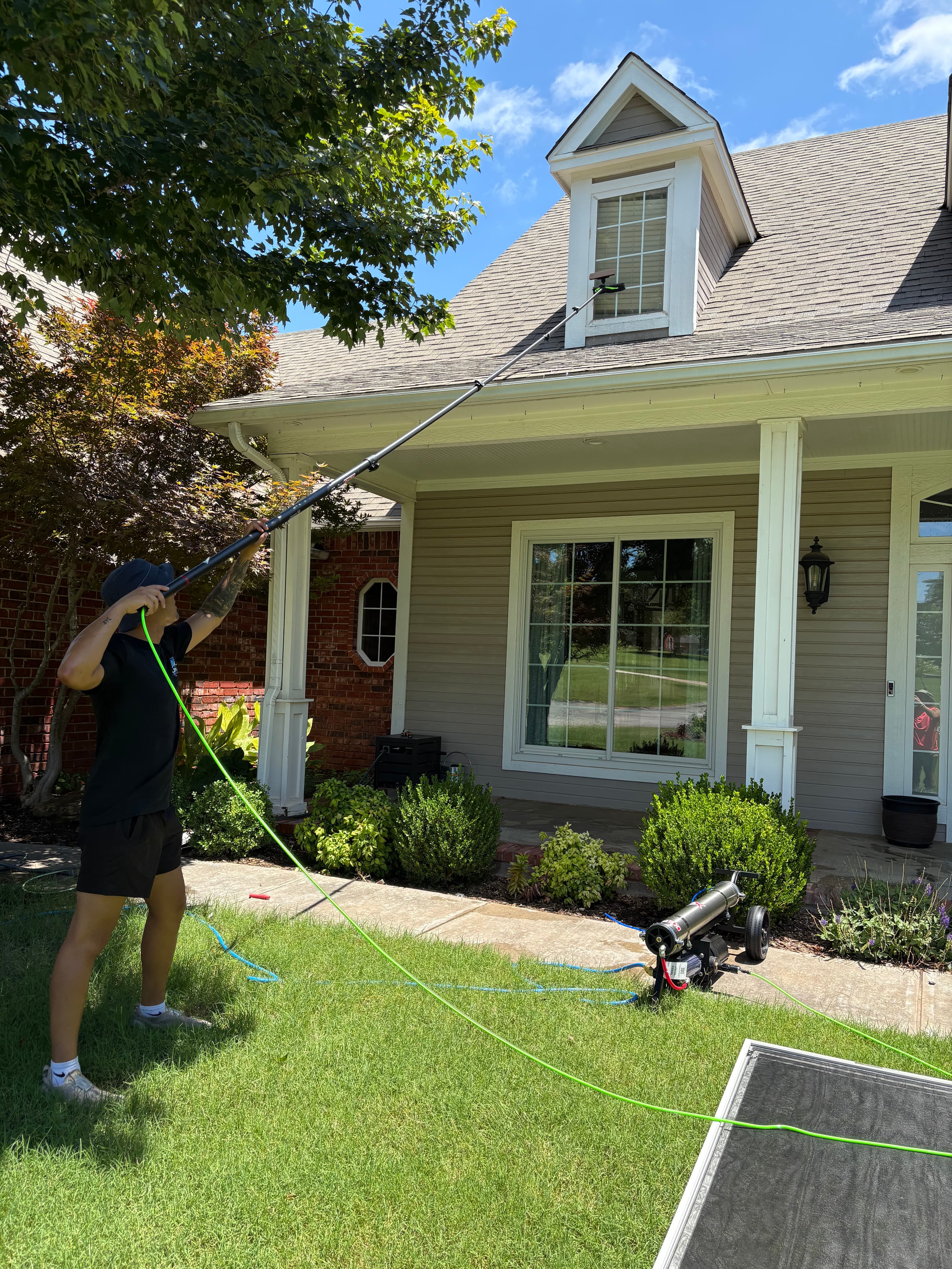 A person cleans a second-story window of a house using a long telescopic water-fed pole.