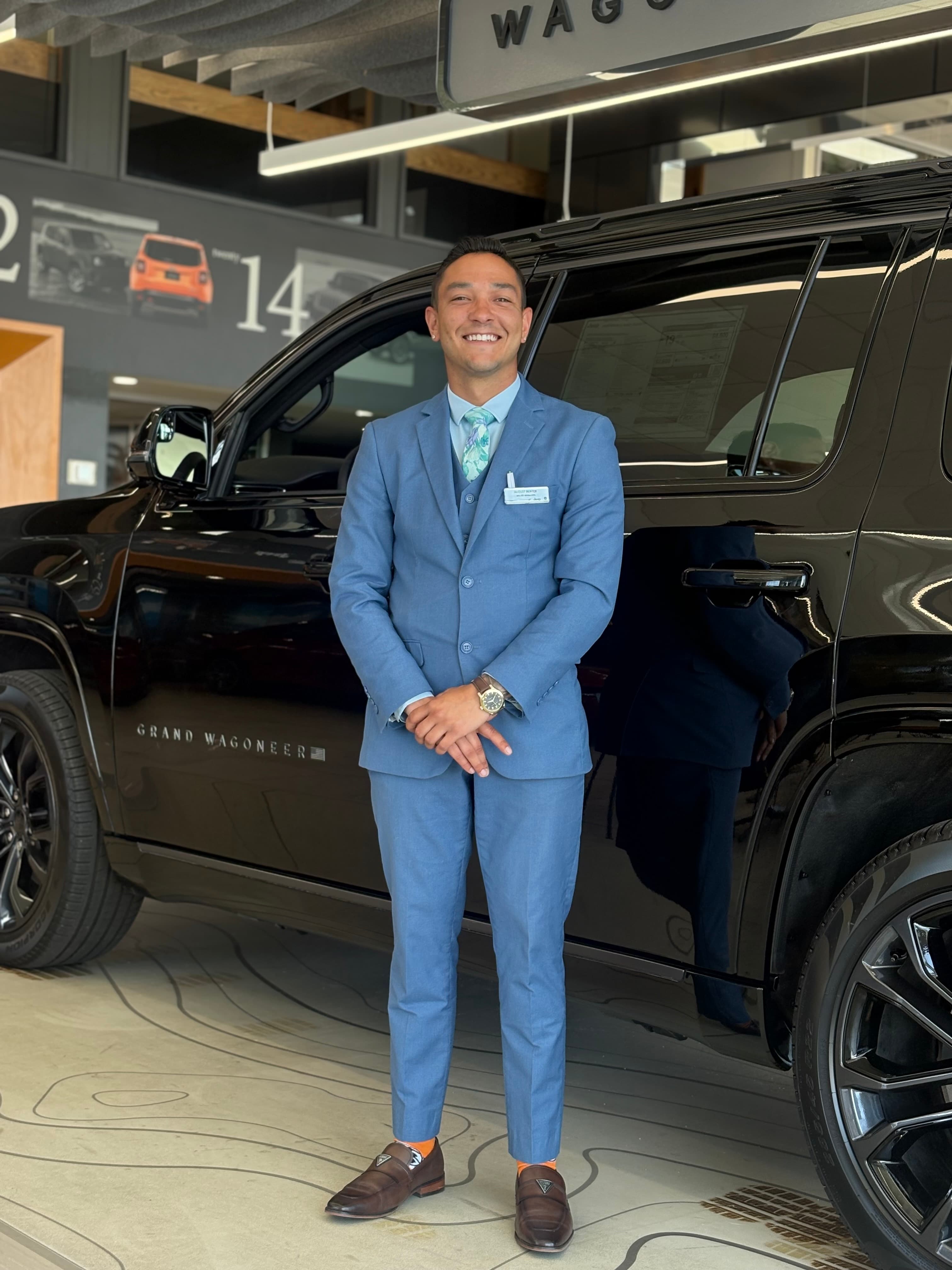 Smiling man in a blue suit standing in front of a black Grand Wagoneer SUV.
