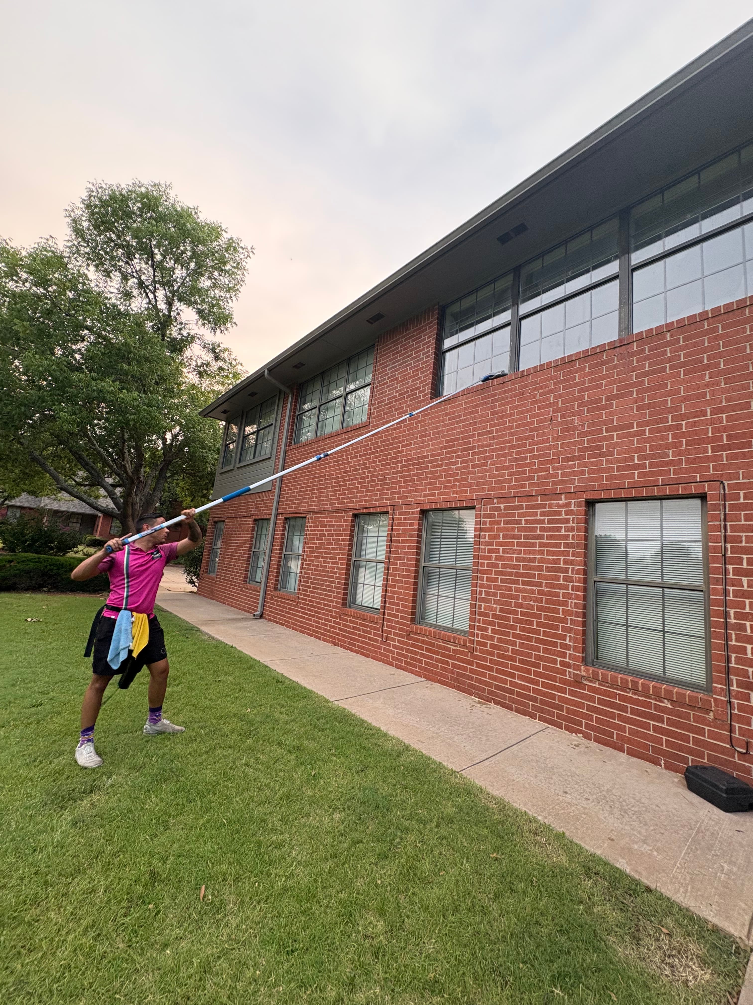 Man cleans second-story windows of a red brick building using a long extension pole.