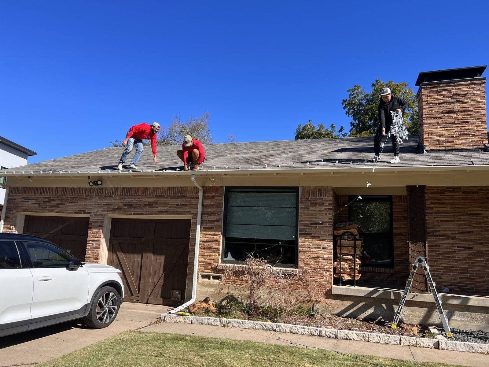 Three men install white Christmas lights along the roofline of a suburban brick house.